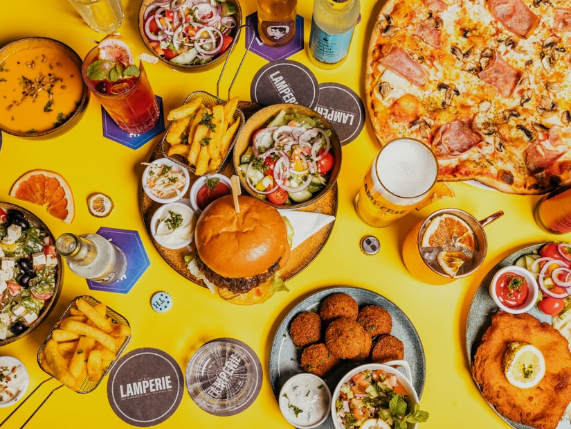 An overhead spread of burgers, pizza, fries, fried snacks and drinks on a bright yellow table