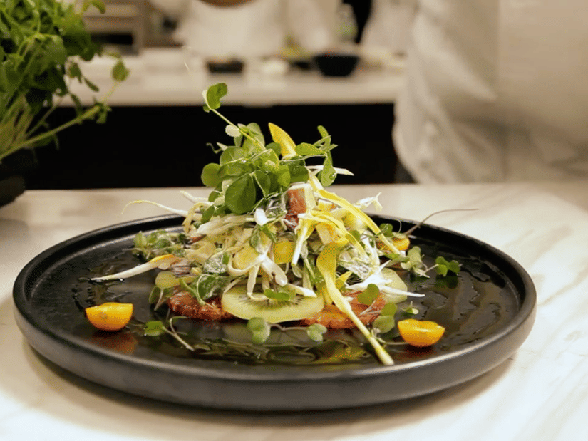 A plated dish of kiwi, shredded vegetables and microgreens on a dark plate on a marble counter
