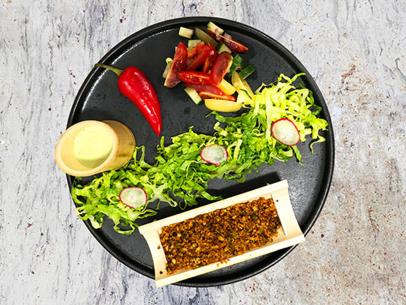 An overhead view of a dark plate with shredded lettuce, radish, red chilli and a wooden tray of spiced grains