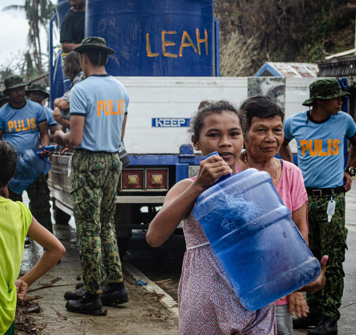 Local police distributing water from a truck while a girl carries a large water container and residents gather nearby after Typhoon Odette