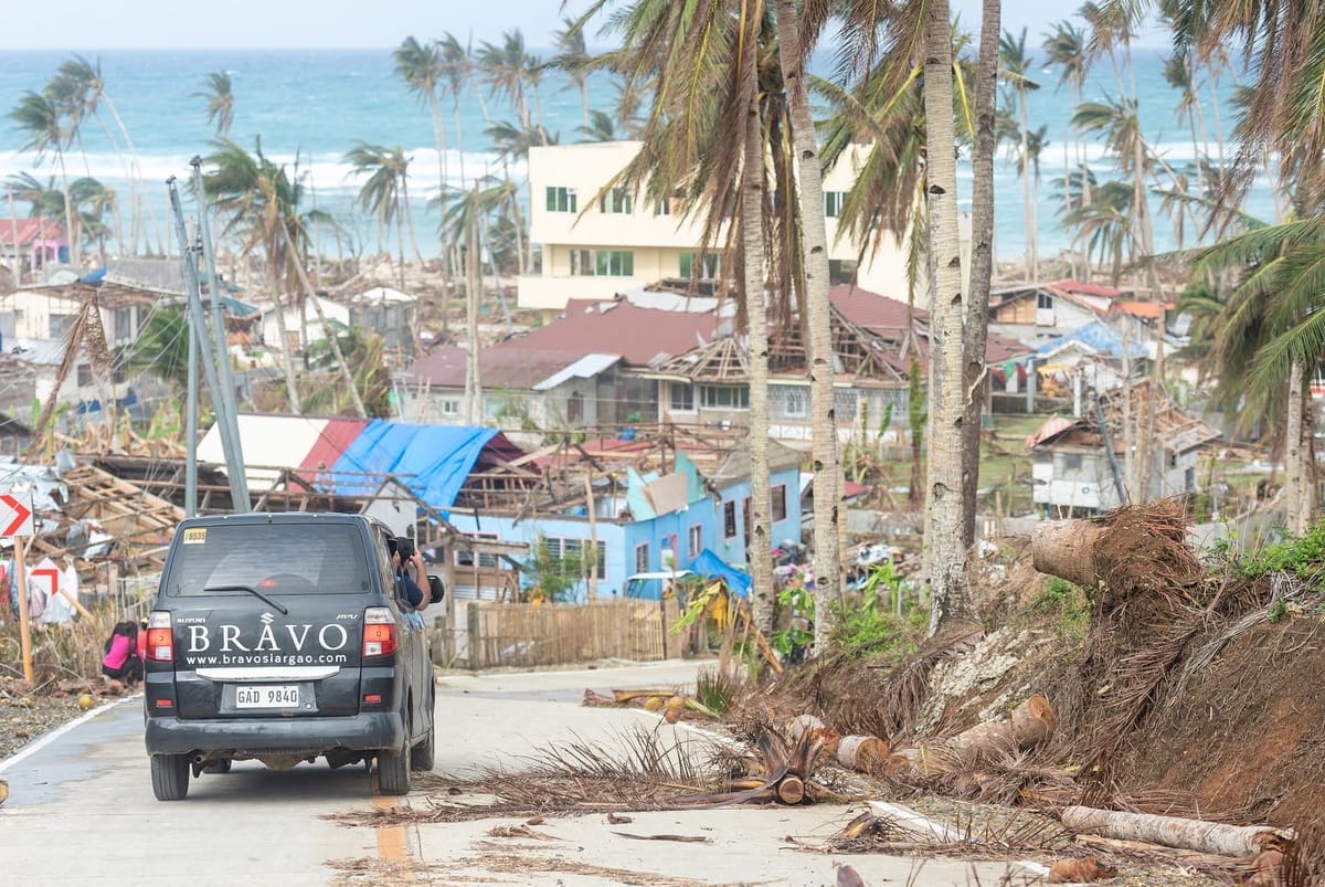 A van branded Bravo Beach Resort Siargao driving along a coastal road through a typhoon-damaged town, with downed palm trees and the ocean beyond