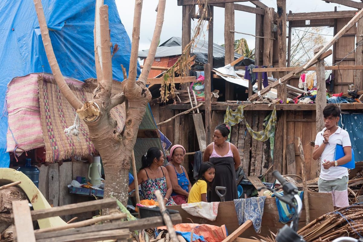 Women and a child seated amid storm wreckage of blue tarps and broken wood beside a stripped tree trunk in Siargao after Typhoon Odette