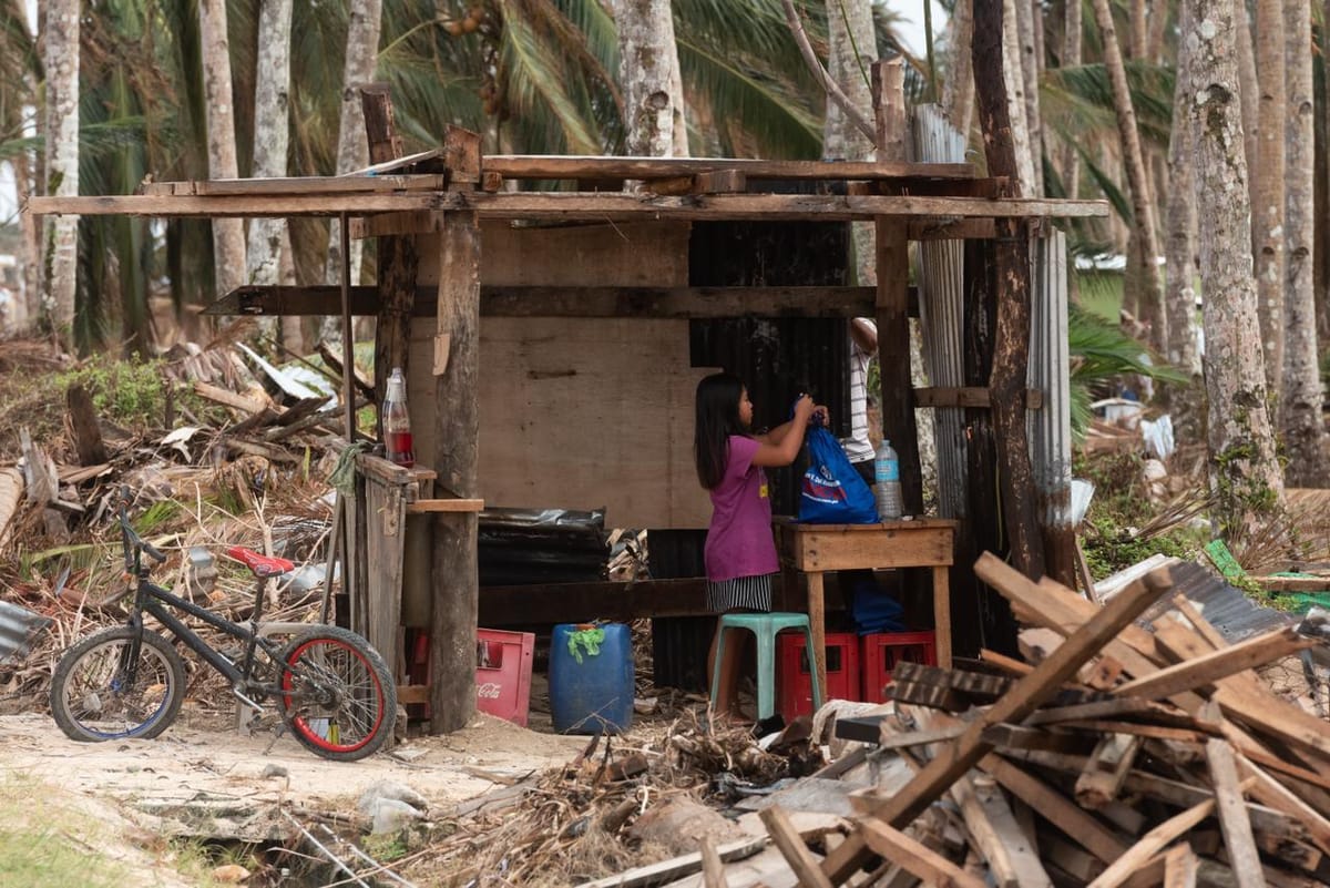 Damaged wooden structure with debris, felled coconut trees, a bicycle, and 2 people salvaging items in the aftermath of a typhoon in Siargao