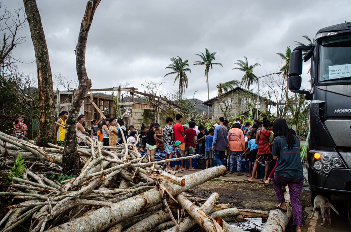 Uprooted trees, storm debris, and damaged houses in Siargao with residents gathered near a truck for cleanup after a typhoon, under a cloudy sky