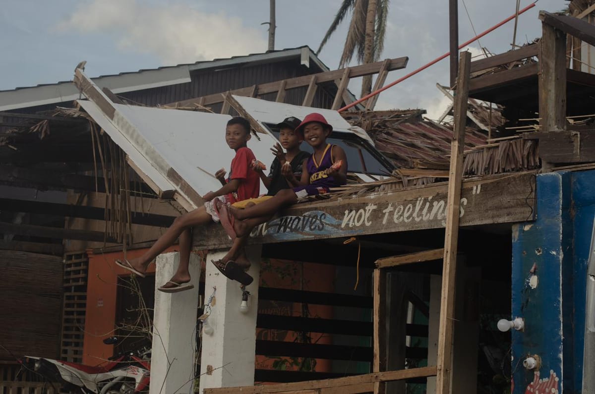 Three children sitting on a storm damaged rooftop with debris scattered around and a wall painted with a waves not feelings message below