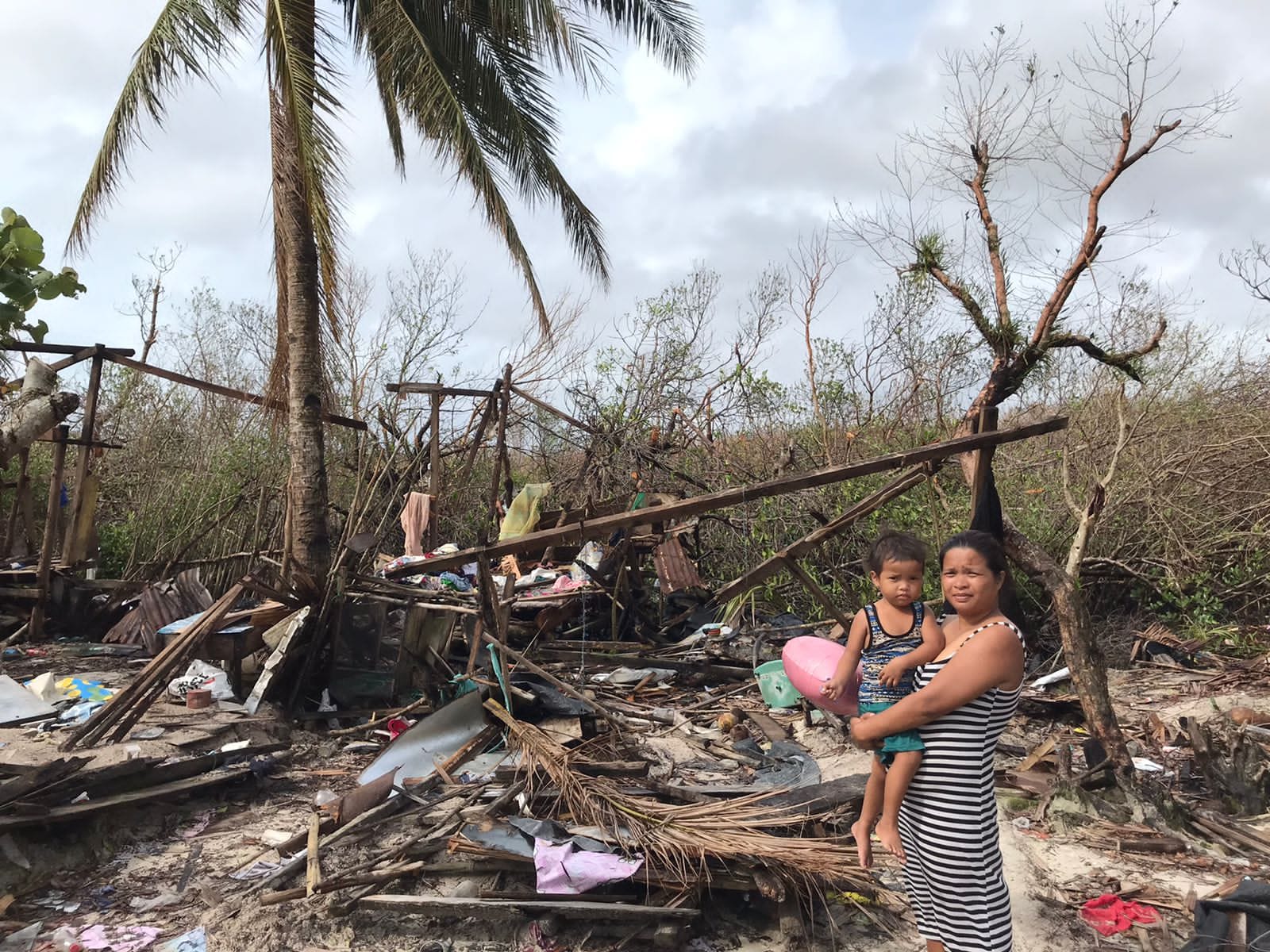 A woman holds a young child while standing amid storm wreckage of broken palm trees and scattered debris after a typhoon in Siargao
