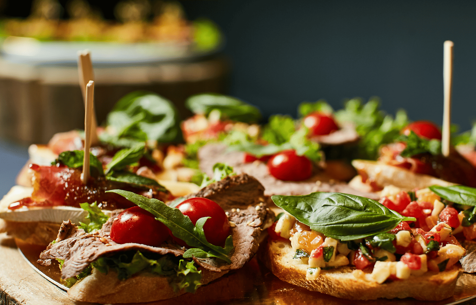 Bruschetta canapes topped with cherry tomatoes, basil, roast beef, and diced vegetables, skewered with toothpicks on a wooden board