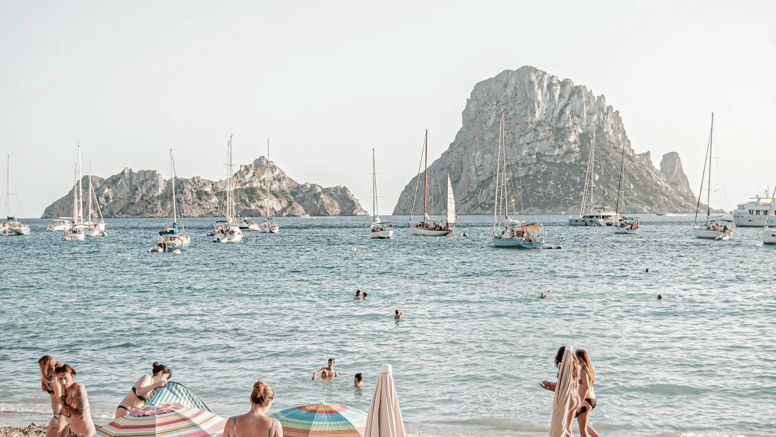 Sailboats anchored near a dramatic rocky island, with beachgoers swimming and relaxing under colorful umbrellas on the sandy shore