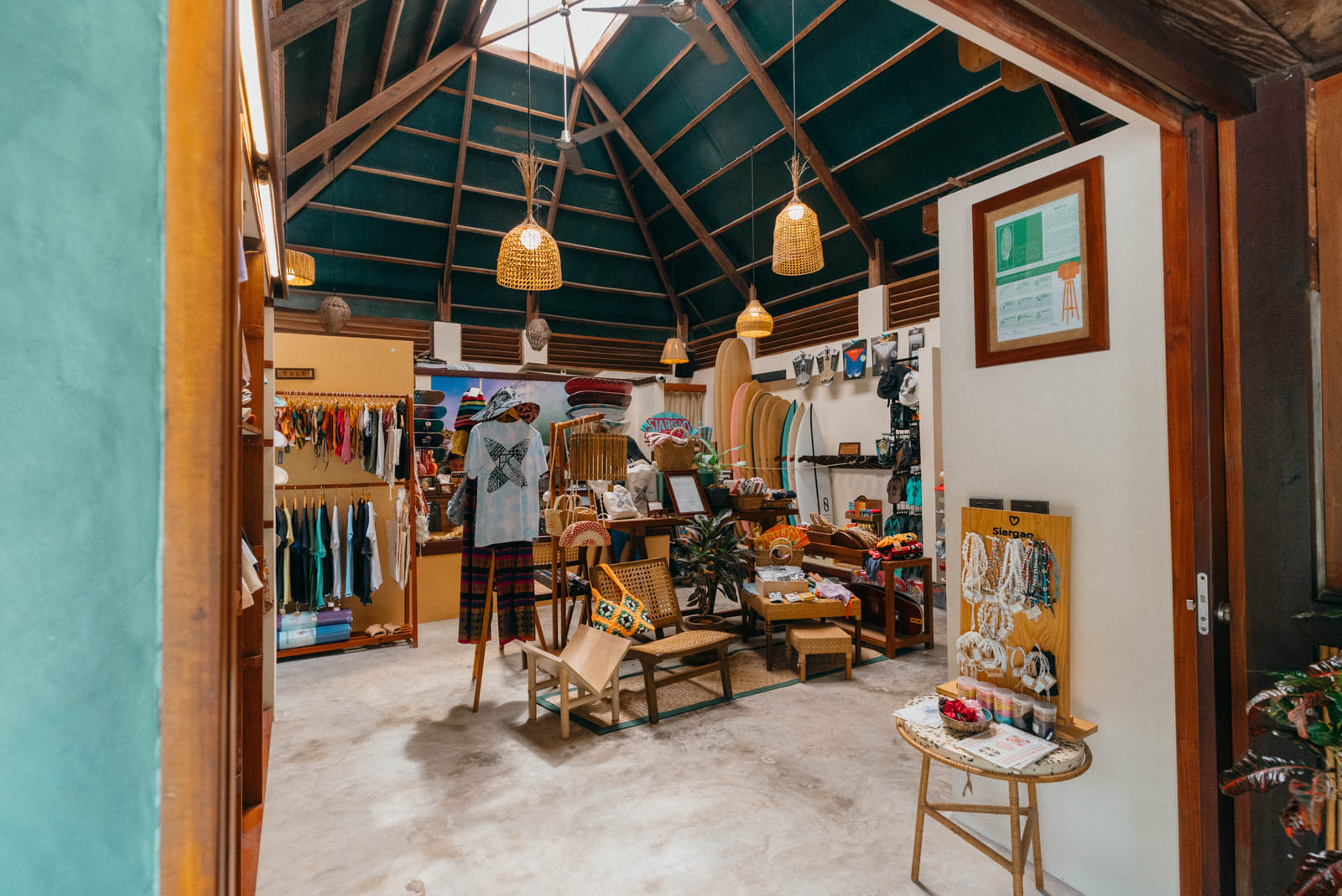 Inside the Bravo Shop at Bravo Beach Resort Siargao, with clothing racks, surfboards, and accessories beneath a high vaulted teal ceiling