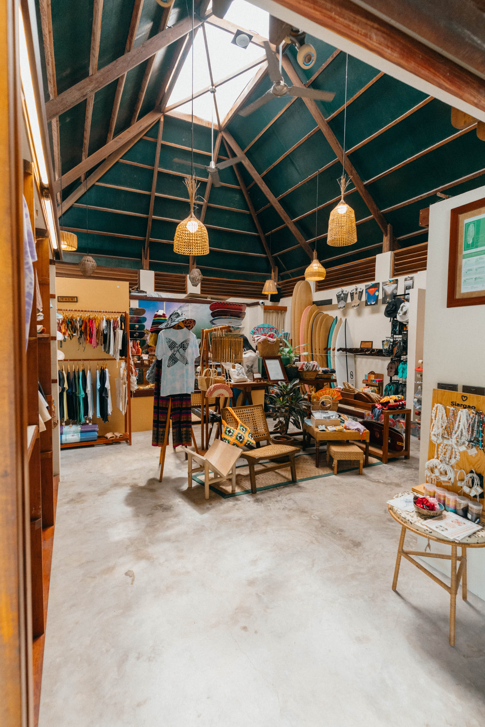 Apparel racks, surfboards, and wicker furniture displayed inside the Bravo Shop at Bravo Beach Resort Siargao, beneath a high teal ceiling with wooden beams