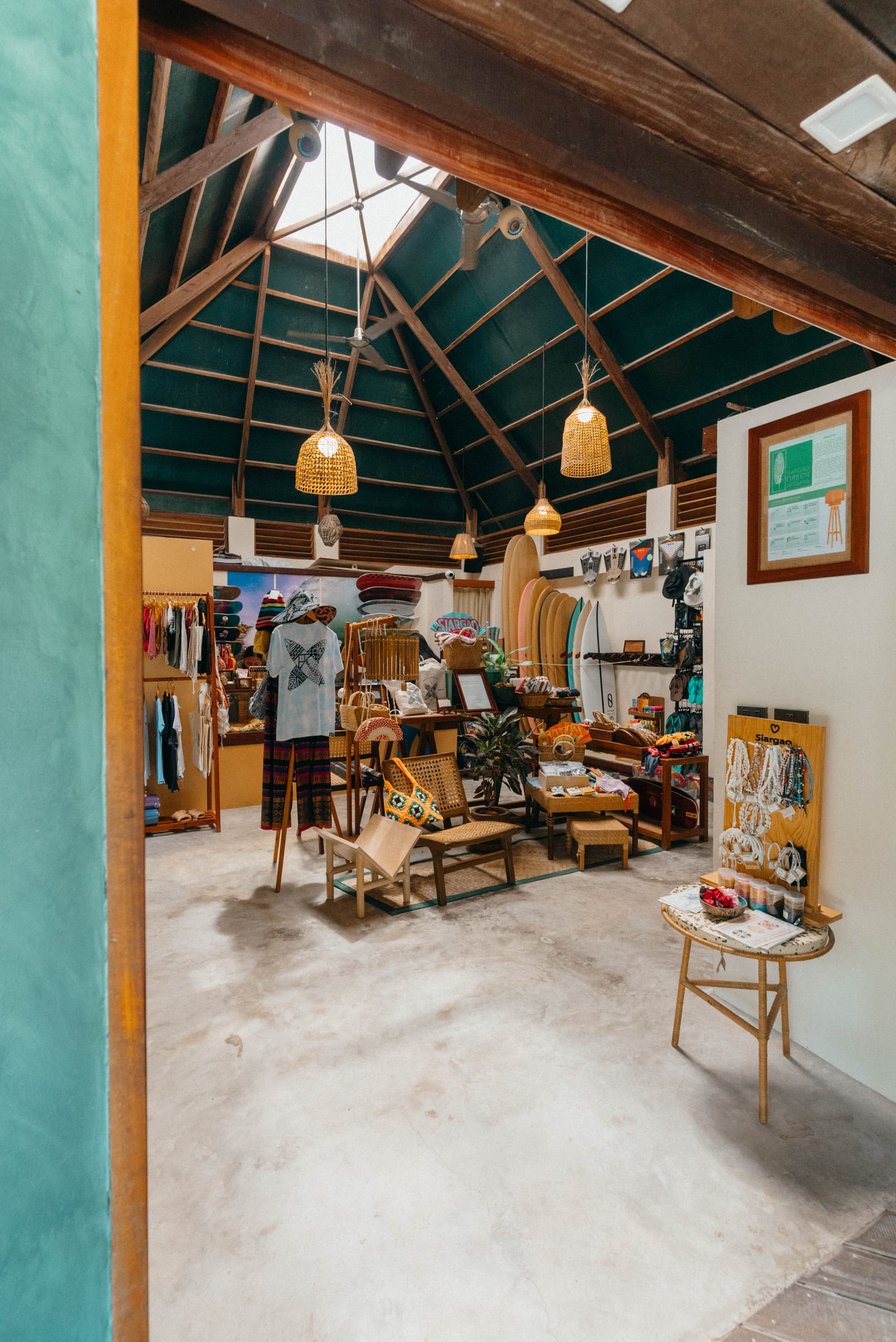 Interior of the Bravo Beach Resort Siargao shop with surfboards, rattan pendant lights, clothing racks, and jewelry displays under a high wooden ceiling