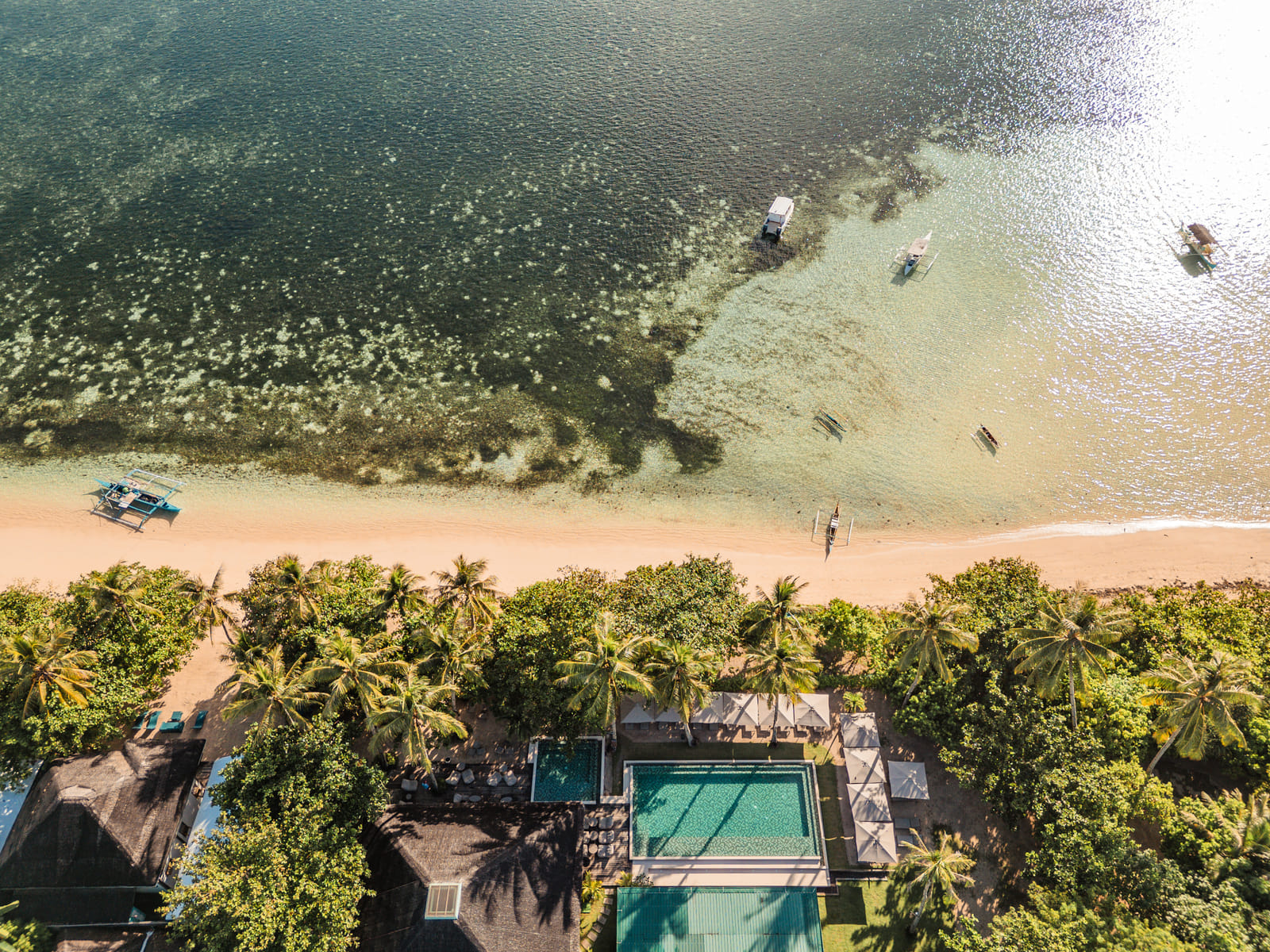 Aerial view of Bravo Beach Resort Siargao, its pools and cabanas set behind the palm-fringed beach with outrigger boats moored in the turquoise shallows