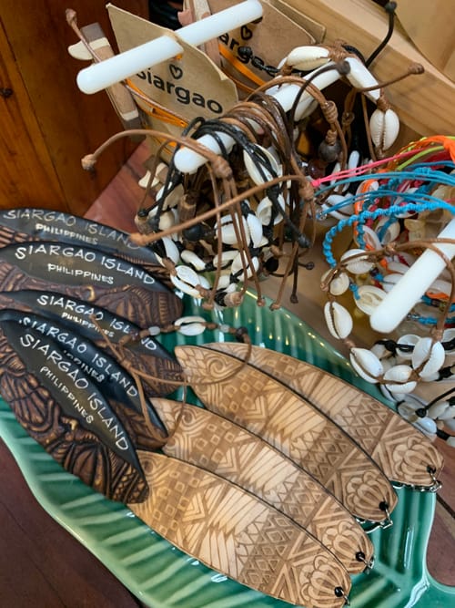 Wooden surfboard-shaped keychains engraved with Siargao Island Philippines, displayed alongside shell and beaded bracelets in a shop