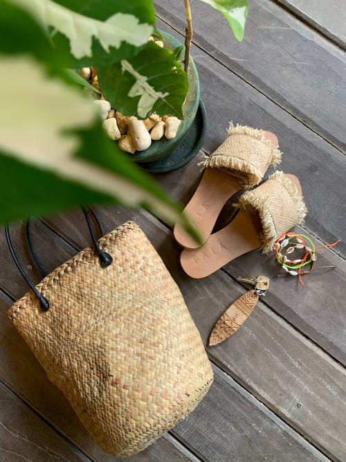 A woven straw bag, a pair of frayed raffia sandals, a set of keys with a wooden keychain, and beaded bracelets arranged on a wooden deck
