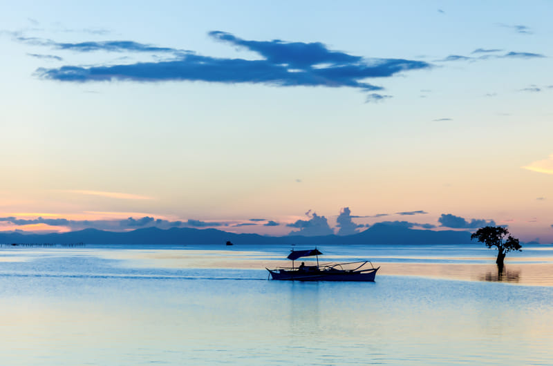 A traditional outrigger boat floats on calm turquoise waters at dusk, with silhouetted mountains and a lone tree along the shoreline