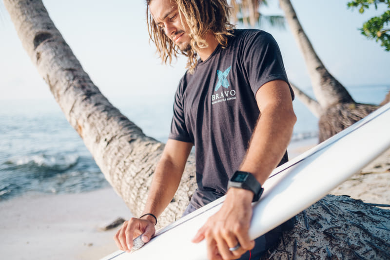 A man with dreadlocks wears a dark grey Bravo Beach Resort Siargao branded t-shirt while waxing a surfboard on the beach beneath coconut palms