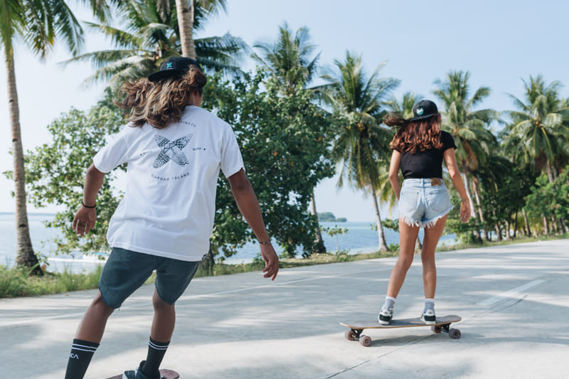 Two people wearing Bravo Beach Resort Siargao branded caps and shirts skateboard along a palm-lined coastal road with the sea in view