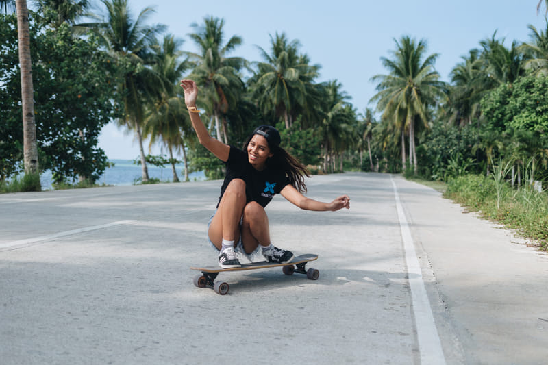 A woman wearing a Bravo Beach Resort Siargao branded t-shirt crouches while skateboarding along a palm-lined road with the sea nearby