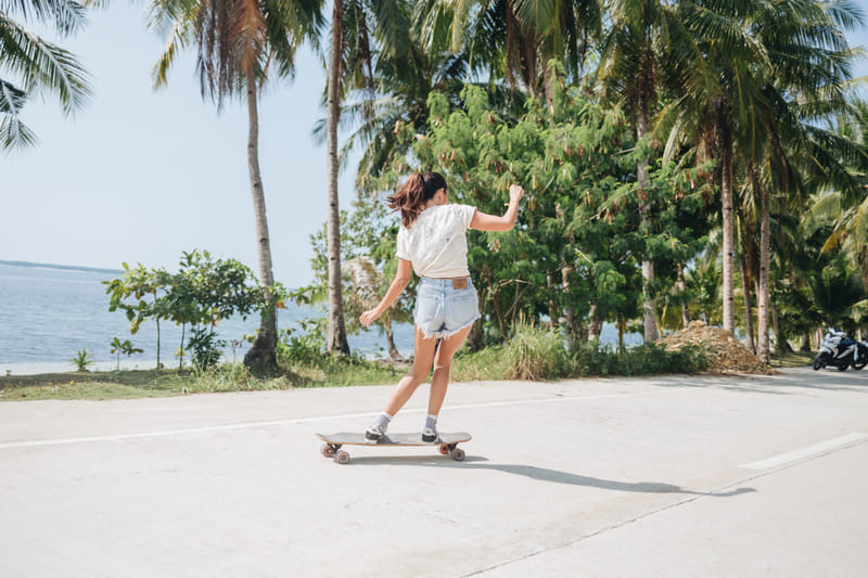 A woman in a white top and denim shorts skateboards along a beachside road lined with coconut palms near Bravo Beach Resort Siargao