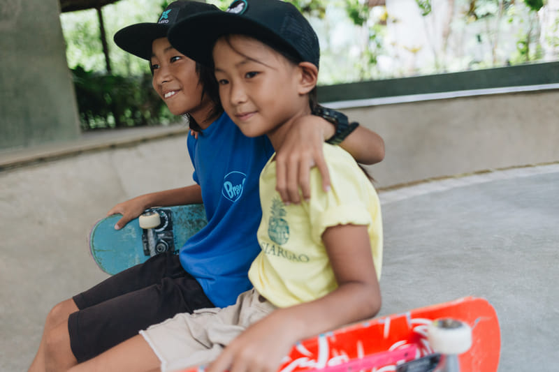 Two children wearing Bravo Beach Resort Siargao branded caps and t-shirts sit together at the skate bowl holding their skateboards
