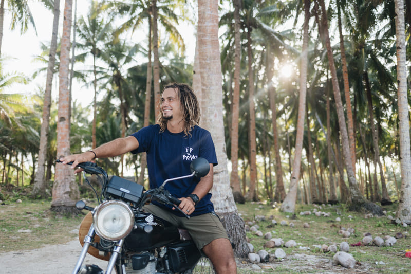 A smiling man with long dreadlocks wearing a Bravo Beach Resort Siargao branded t-shirt sits on a motorcycle in a sunlit coconut grove