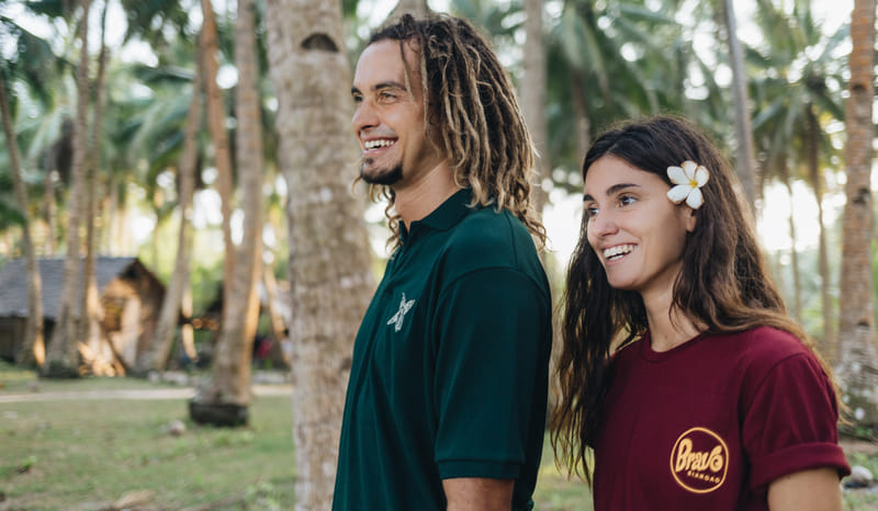 A smiling man and woman wearing Bravo Beach Resort Siargao branded shirts stand together in a coconut grove near a thatched hut roof