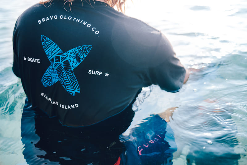 Back view of a black Bravo Clothing Co t-shirt with a crossed surfboard graphic and Skate and Surf text, worn while standing in shallow water