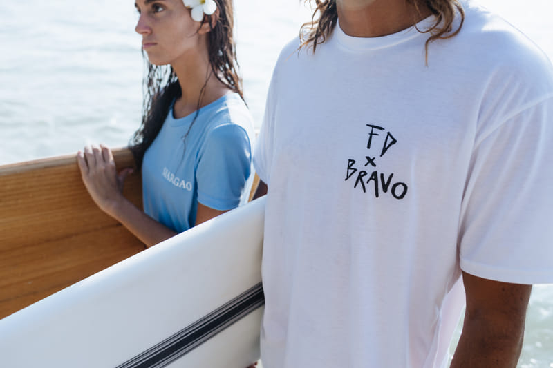 A man wearing a white Bravo Beach Resort Siargao branded t-shirt holds a surfboard beside a woman with a flower in her hair on a boat