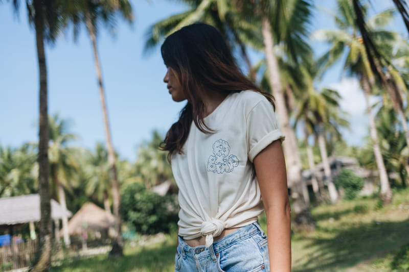 A woman wearing a cream t-shirt with a floral graphic, knotted at the waist, walks through a coconut grove near thatched huts at Bravo Beach Resort Siargao