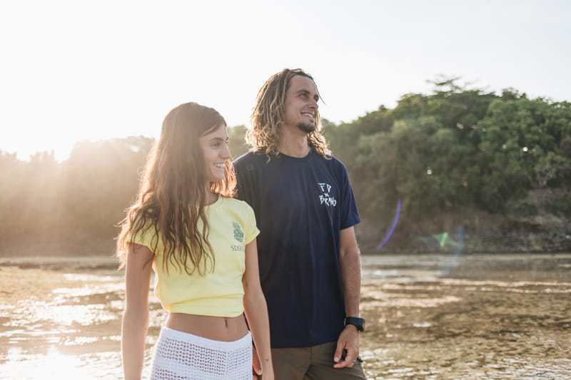 A man wearing a navy Bravo Beach Resort Siargao branded t-shirt and a woman in a yellow top stand together at a lagoon shoreline at sunset