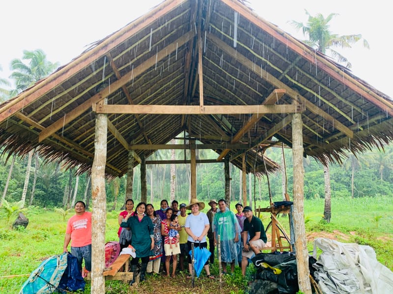 Group of people standing under an open thatched-roof farm shelter in the rain, surrounded by palm trees and fields