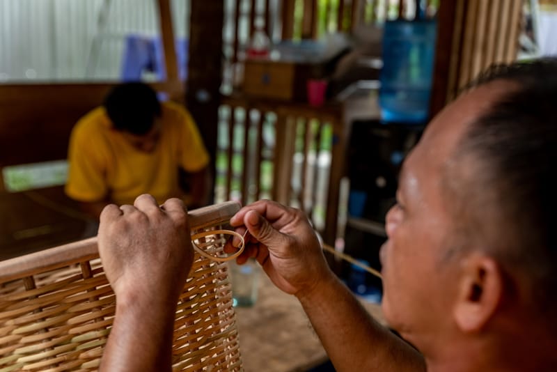 Craftsman weaving rattan strands onto a wooden chair frame by hand, another worker in a yellow shirt visible at a Siargao Green Artisans workshop