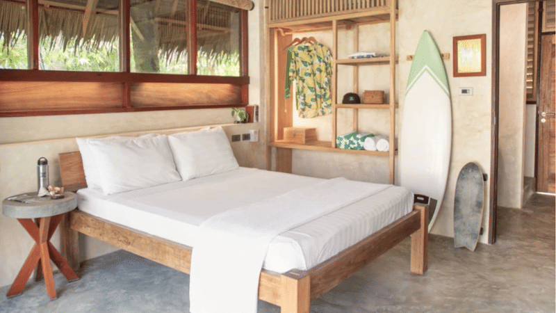 Bravo Beach Resort Siargao guest room with a wooden bed, white linens, open wardrobe shelving, and a surfboard leaning against the wall