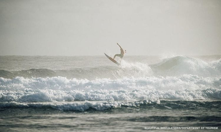 A surfer launches off the top of a breaking wave amid choppy white water, credited to Beautiful Destinations and Department of Tourism
