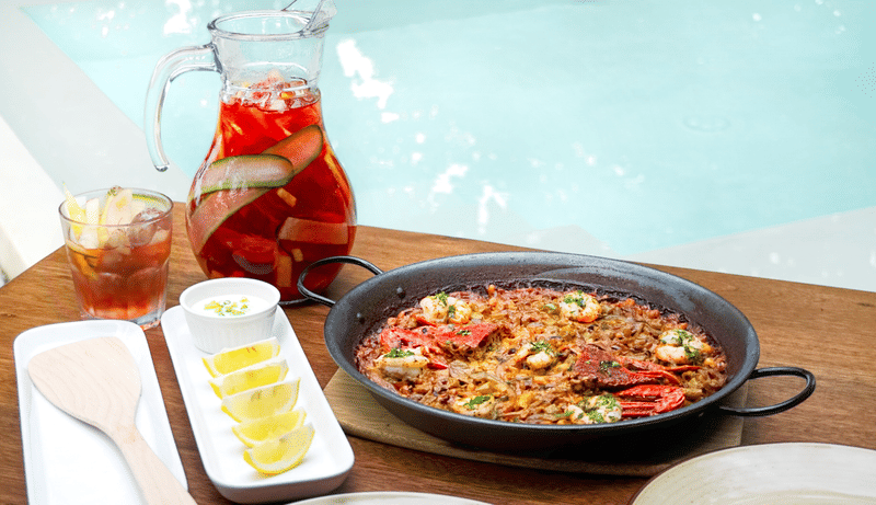 Seafood paella platter with a pitcher of sangria and lemon wedges on a wooden table beside the pool at Bravo Beach Resort Siargao