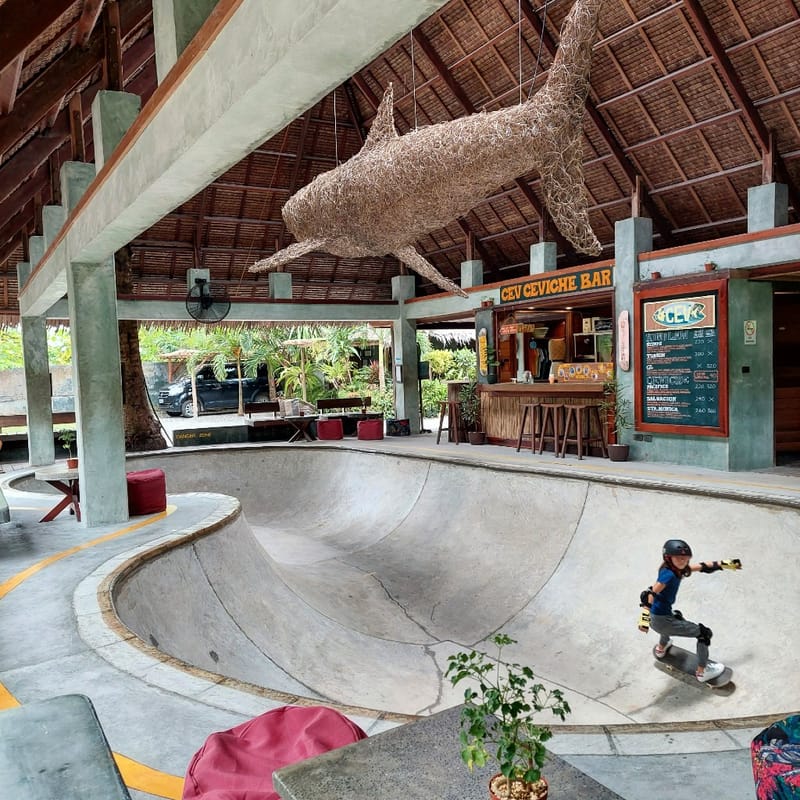 A child wearing a helmet and pads skateboards in the concrete bowl at Bravo Beach Resort Siargao, near the Cev Ceviche Bar and shark sculpture