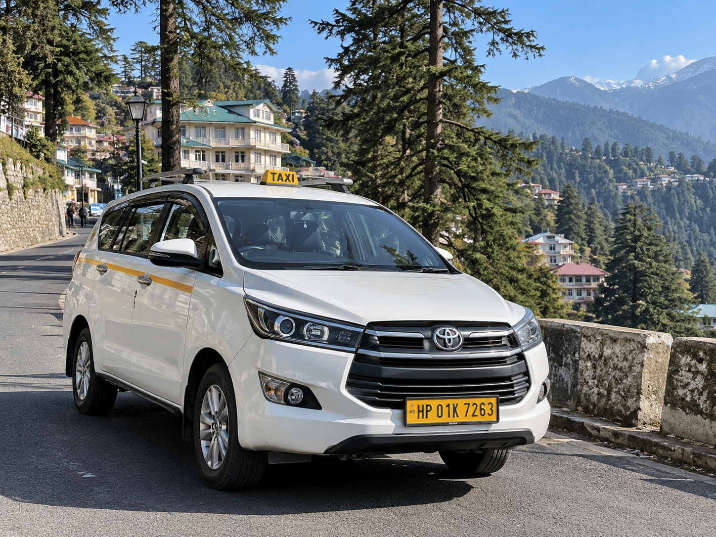 A white Toyota Innova Crysta taxi on a pine-lined hill road in Himachal Pradesh