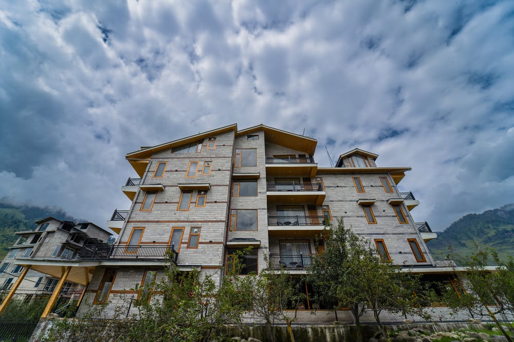 Low-angle view of Nazaara Luxe Retreat's facade against a cloud-streaked mountain sky, apple orchard in the foreground