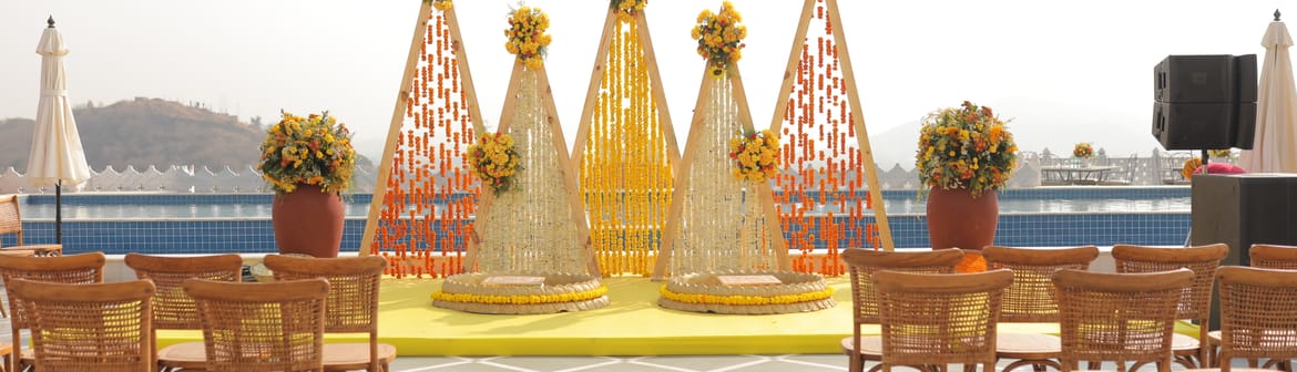 Conical marigold backdrops on a yellow stage with rows of cane chairs on a poolside terrace, white parasols, lake and hills beyond, at Fateh Vilas, Udaipur.