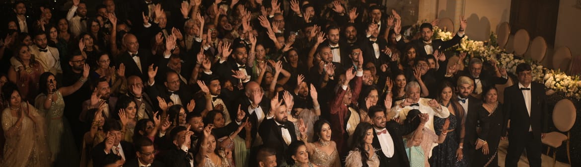 Guests in formal wear raising their hands in the Surya Prakash Hall, with carved stone pillars, arched doors and strung lights at Fateh Vilas, Udaipur.