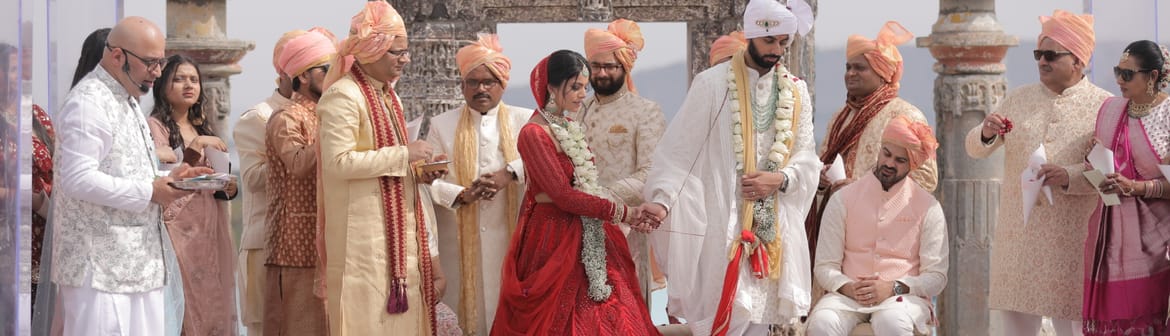 Bride in red and groom in white stand hand in hand before a carved stone gateway, family gathered on a petal-strewn floor at Fateh Vilas, Udaipur.