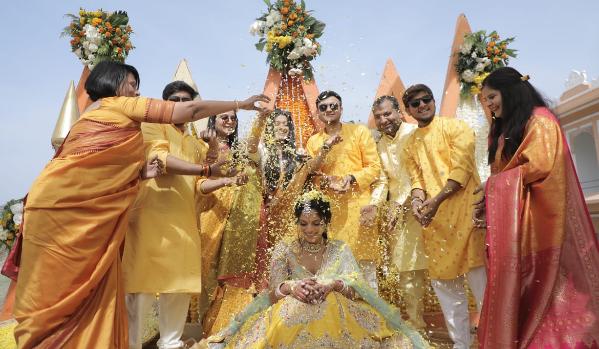 Bride seated in a yellow lehenga as family in yellow and orange shower her with petals, conical marigold backdrops behind, at Fateh Vilas, Udaipur.