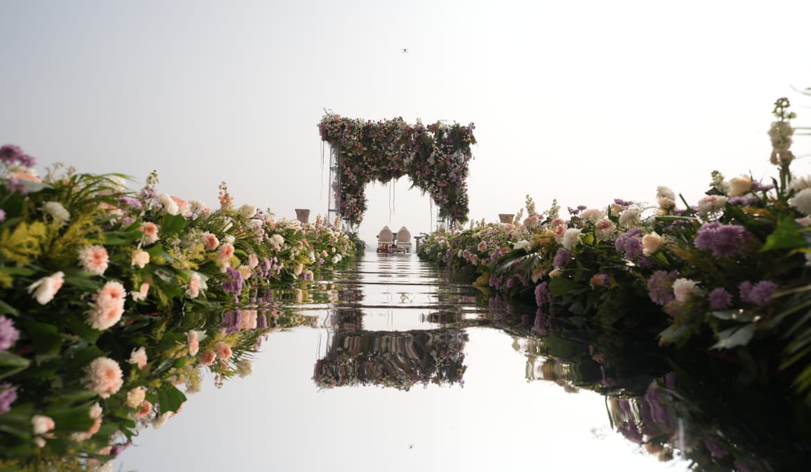 Floral mandap at the end of a mirrored walkway, flanked by banks of pastel flowers and reflected in the water below under a misty sky at Fateh Vilas, Udaipur.