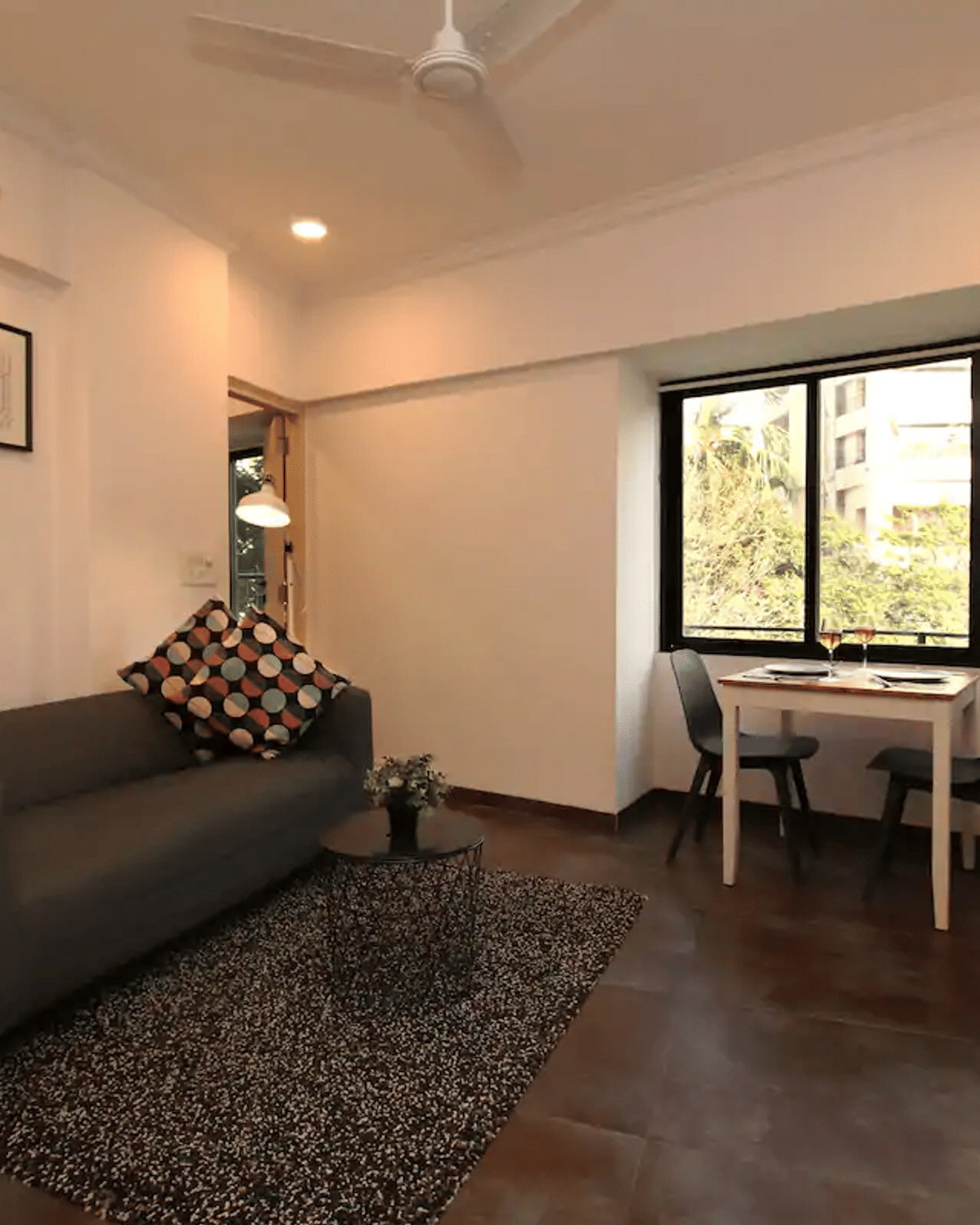 Living room of the 1BHK apartment at Wabi-Sabi, Juhu, with a grey sofa, framed art, a TV console, and a 2-seater dining table beside a window with wine glasses.