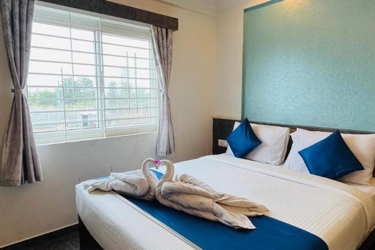 Queen bed with white linen, blue cushions, and a towel swan in the Standard AC Room at Value Inn, Bangalore Airport beside a bright window