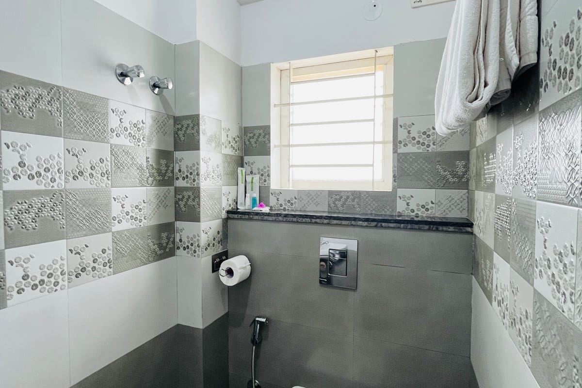 Guest bathroom at Value Inn, Bangalore Airport with grey and white wall tiles, a bright window, hanging towels, and a wall mounted toilet