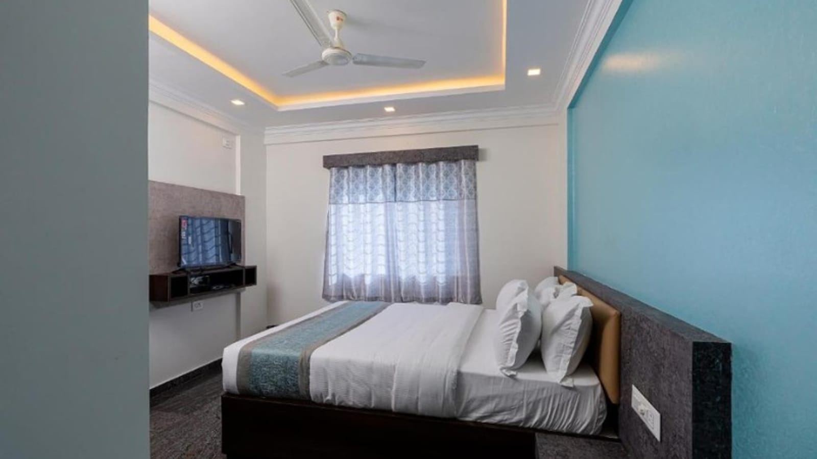 Standard Non-AC Room at Value Inn, Bangalore Airport with a queen bed, teal feature wall, ceiling fan, cove lighting, and a bright window