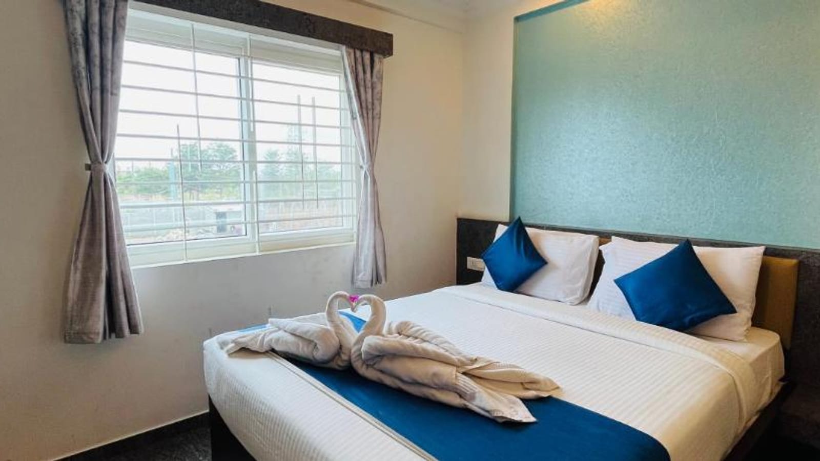Queen bed with white linen, blue cushions, and a towel swan in the Standard Non-AC Room at Value Inn, Bangalore Airport by the window