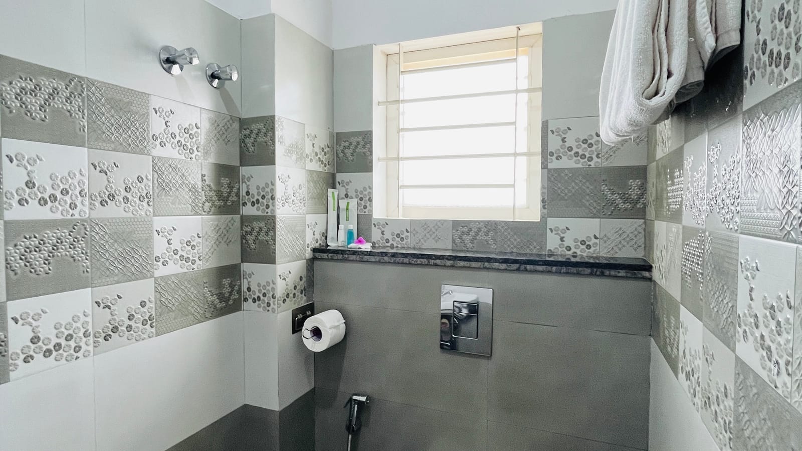 Guest bathroom at Value Inn, Bangalore Airport with grey and white wall tiles, a bright window, hanging towels, and a wall mounted toilet