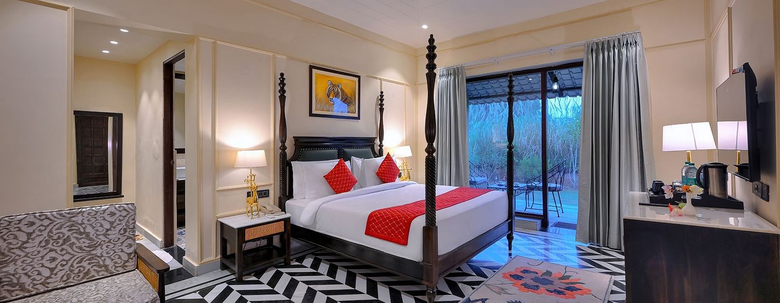 Premium Suite Cottage featuring a four-poster bed with red accents, chevron flooring, patterned sofas and a coffee table in the seating area, a TV, and a balcony with chairs - Singhvilas, Ranthambore